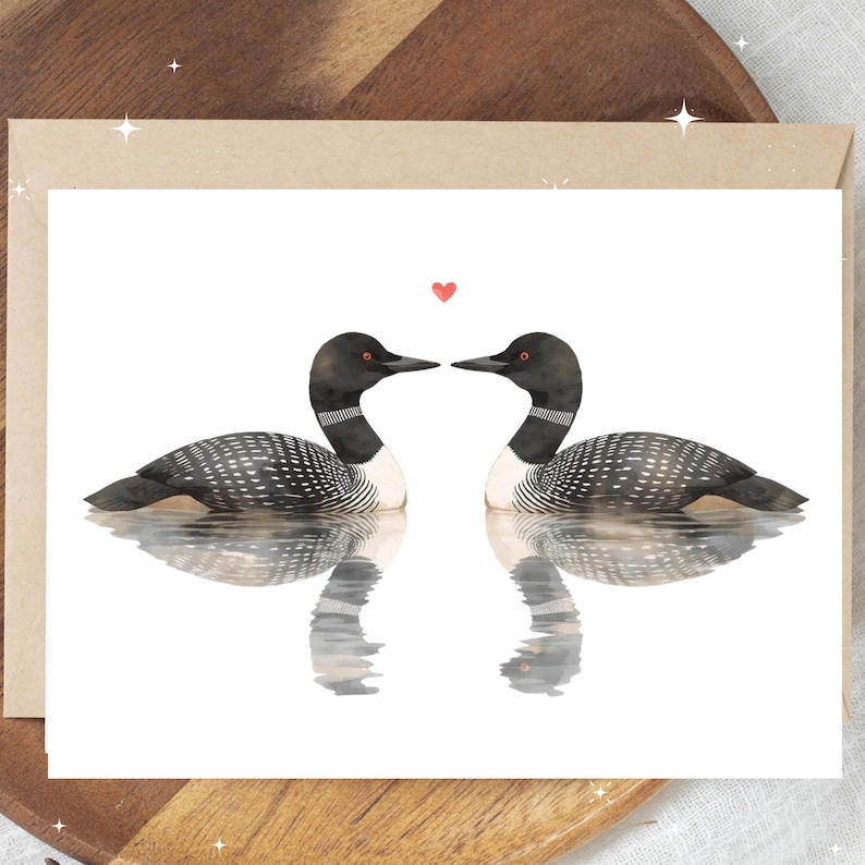 May include: A greeting card featuring two loons facing each other, with a small red heart above them. The loons are depicted in a watercolor style, with black and white speckled feathers and black heads. The card is on a white background.