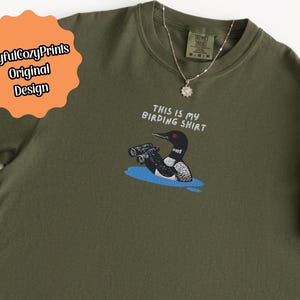 May include: Olive green t-shirt with the text "THIS IS MY BIRDING SHIRT" and a graphic of a bird with binoculars. A silver necklace with a sun pendant is on the shirt. The shirt has a tag that says "Brand: Brand Name" and "M-M-M".