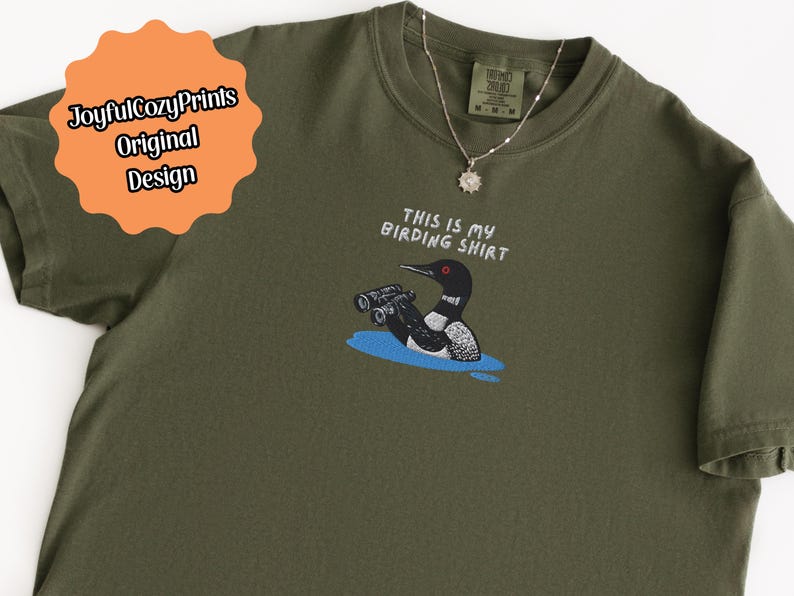 May include: Olive green t-shirt with the text "THIS IS MY BIRDING SHIRT" and a graphic of a bird with binoculars. A silver necklace with a sun pendant is on the shirt. The shirt has a tag that says "Brand: Brand Name" and "M-M-M".