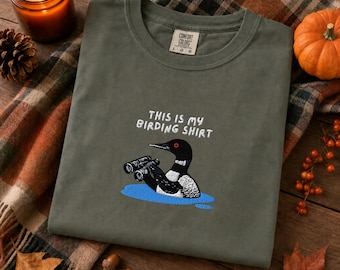 Könnte beinhalten: Ein olivgrünes T-Shirt mit Rundhalsausschnitt und einer Stickerei eines Eistauchers, der ein Fernglas in blauem Wasser hält. Der Text über dem Vogel lautet "THIS IS MY BIRDING SHIRT". Das T-Shirt hat ein Comfort Colors-Etikett am Innenkragen.