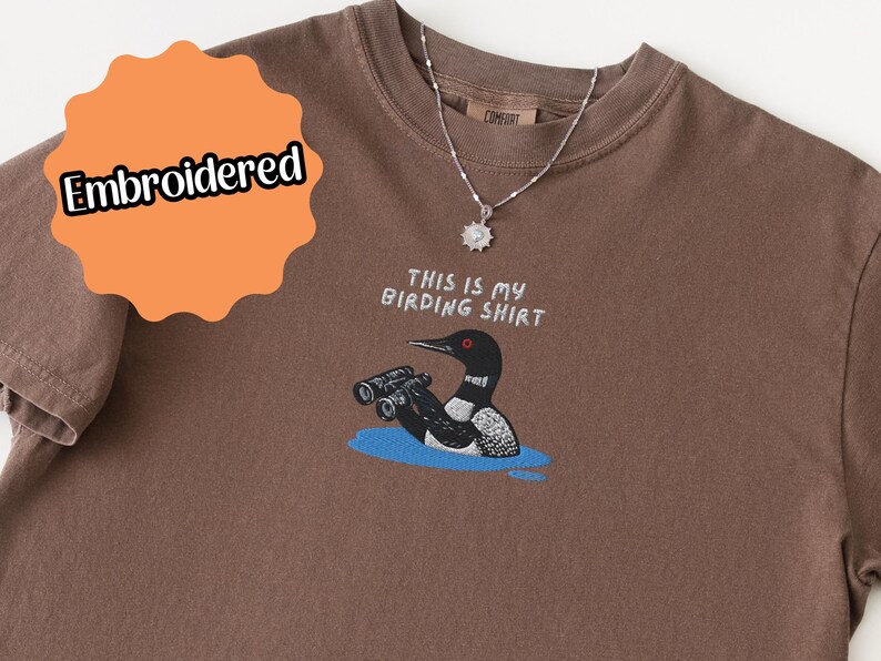 May include: Brown t-shirt with an embroidered design. The design features a loon with binoculars in a blue water setting, and the text "THIS IS MY BIRDING SHIRT". A silver necklace with a pendant is also visible.