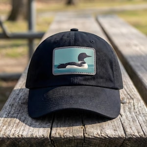 May include: Black baseball cap featuring a rectangular patch with a graphic of a loon swimming in water. The loon is black and white with a black head and beak. The background is a light blue color.
