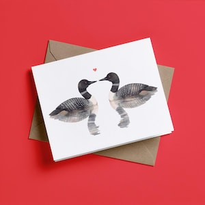 May include: A white greeting card featuring a watercolor illustration of two loons facing each other on water, with a small red heart positioned between their beaks. The card is placed on top of a brown envelope.