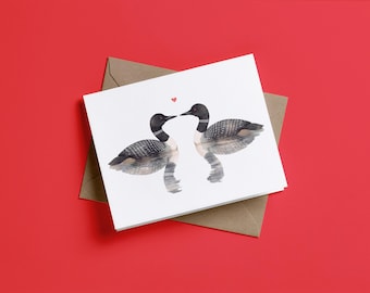 May include: A white greeting card featuring a watercolor illustration of two loons facing each other on water, with a small red heart positioned between their beaks. The card is placed on top of a brown envelope.