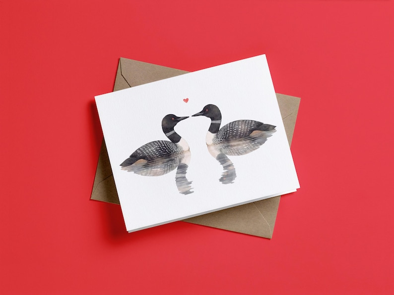 May include: A white greeting card featuring a watercolor illustration of two loons facing each other on water, with a small red heart positioned between their beaks. The card is placed on top of a brown envelope.
