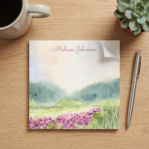 Peut inclure: Bloc-notes avec un paysage à l'aquarelle représentant des fleurs violettes, de l'herbe verte et des collines bleu-vert sous un ciel doux et clair. Le nom "Melissa Johnson" est écrit en cursive en haut.