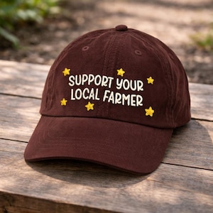 May include: A brown corduroy baseball cap with the embroidered text "SUPPORT YOUR LOCAL FARMER" in white, accented by yellow star designs. The cap is sitting on a wooden surface.