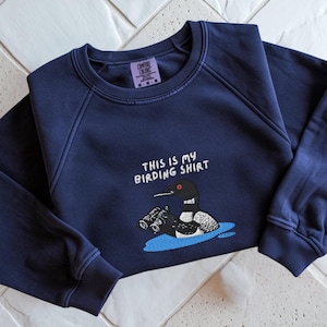 May include: Navy blue sweatshirt with the text "THIS IS MY BIRDING SHIRT" above an embroidered cartoon loon holding binoculars. The loon is in a blue water scene. The sweatshirt has a crew neck and long sleeves.