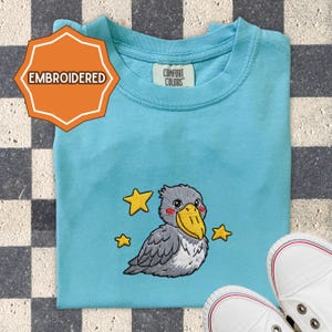 May include: A light blue t-shirt with an embroidered design of a cartoon bird with a yellow beak and three yellow stars. The shirt has the word "EMBROIDERED" on a tag. The brand tag says "COMFORT COLORS". White sneakers are in the bottom right corner.