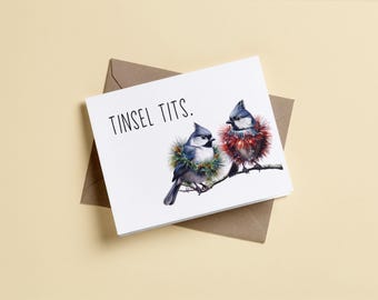 May include: A white greeting card featuring two blue and grey birds perched on a branch, one wearing a green tinsel wreath and the other a red tinsel wreath, with the text TINSEL TITS. printed in black above the birds, resting on a brown envelope.