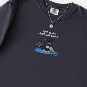 May include: Dark navy blue t-shirt with the text "THIS IS MY BIRDING SHIRT" and a cartoon illustration of a loon with binoculars. A silver necklace with a pendant is also visible. The shirt is made of a soft material.