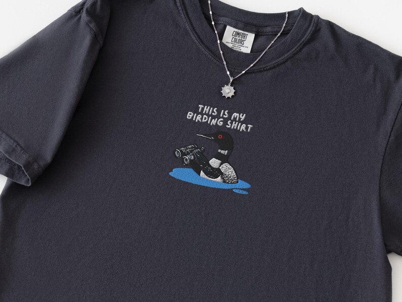 May include: Dark navy blue t-shirt with the text "THIS IS MY BIRDING SHIRT" and a cartoon illustration of a loon with binoculars. A silver necklace with a pendant is also visible. The shirt is made of a soft material.