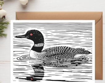 May include: A white greeting card featuring a black and white linocut illustration of a loon swimming on water. The bird has a distinct red eye and detailed feather patterns, with its reflection visible in the water below the surface.