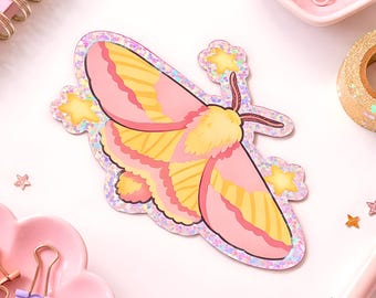 May include: A holographic sticker featuring a moth with pink and yellow wings, a fuzzy yellow body, and brown antennae. The moth is surrounded by small yellow star shapes, all set against a shimmering, iridescent, glitter-like background.