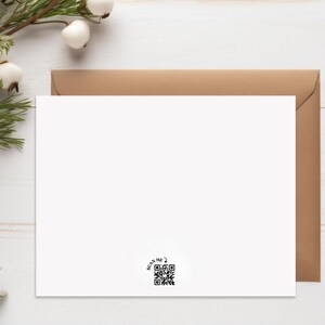 May include: A white greeting card with a brown envelope. The card has a QR code in the lower right corner with the text "SCAN ME?". The card is on a white wooden surface with greenery and white berries in the upper left corner.