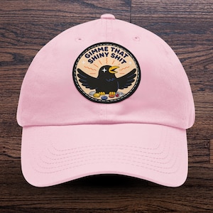 May include: A light pink baseball cap featuring a circular patch with a black crow illustration. The crow has its wings spread and is surrounded by small colorful objects. The text GIMME THAT SHINY SHIT is printed in dark blue letters above the bird.
