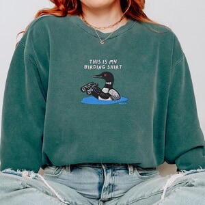 May include: A teal-colored sweatshirt with the text "THIS IS MY BIRDING SHIRT" above an illustration of a loon holding binoculars in a blue pond. The sweatshirt is a relaxed fit.