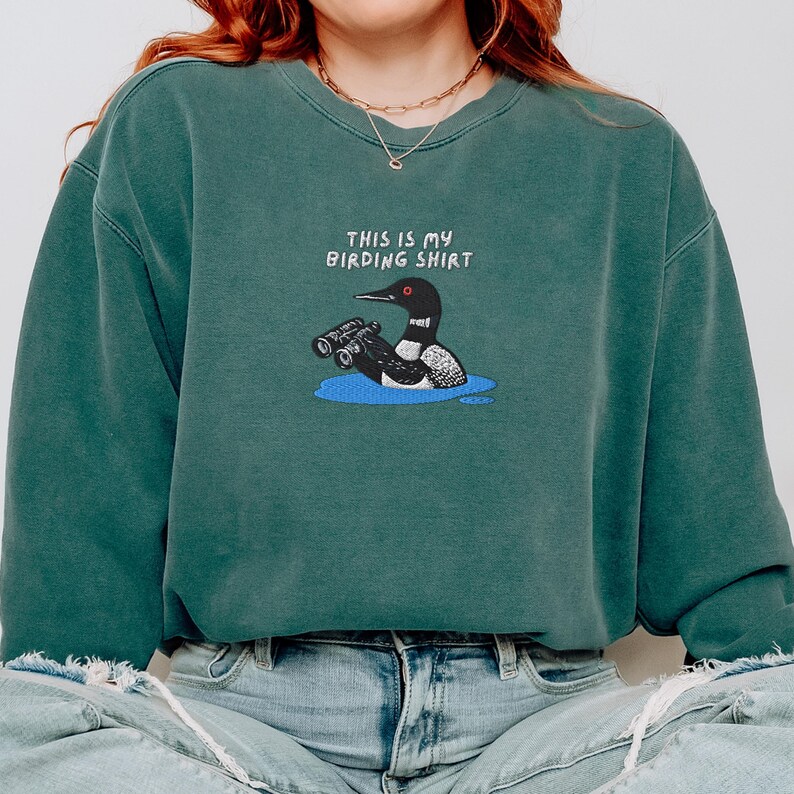 May include: A teal-colored sweatshirt with the text "THIS IS MY BIRDING SHIRT" above an illustration of a loon holding binoculars in a blue pond. The sweatshirt is a relaxed fit.