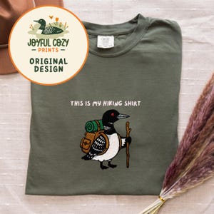 May include: Olive green t-shirt with a cartoon loon carrying a backpack and a walking stick. Text above the loon reads "THIS IS MY HIKING SHIRT." A circular logo in the upper left corner says "JOYFUL COZY - PRINTS - ORIGINAL DESIGN."