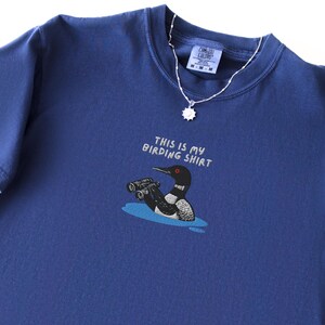May include: A navy blue t-shirt with the text "THIS IS MY BIRDING SHIRT" above a cartoon of a loon with binoculars. A silver necklace with a sun-shaped pendant is draped over the shirt.