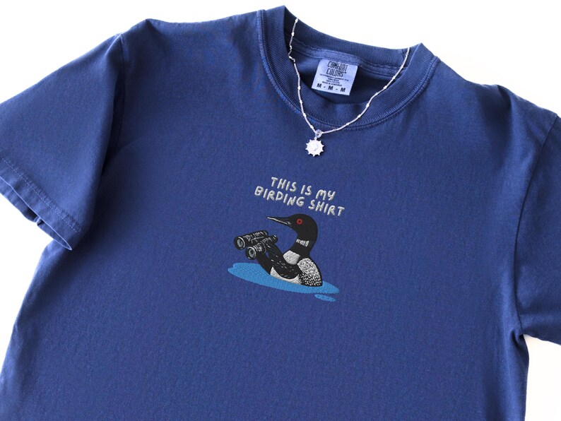 May include: A navy blue t-shirt with the text "THIS IS MY BIRDING SHIRT" above a cartoon of a loon with binoculars. A silver necklace with a sun-shaped pendant is draped over the shirt.