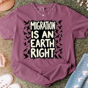 May include: A mauve t-shirt featuring the text "MIGRATION IS AN EARTH RIGHT" in a bold, white font. The design includes black silhouettes of birds in flight, creating a visual theme of movement and freedom. A delicate necklace is visible.