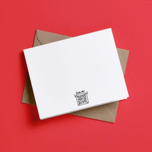 May include: A white rectangular greeting card resting on a brown envelope. The card features a small black printed QR code at the bottom center with the text "SCAN ME!" written above it in a simple, hand-drawn style font.