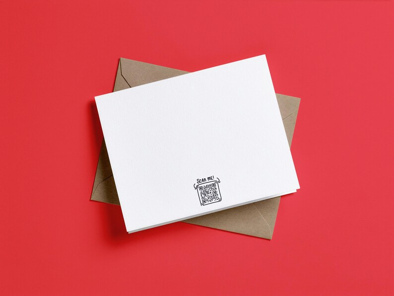May include: A white rectangular greeting card resting on a brown envelope. The card features a small black printed QR code at the bottom center with the text "SCAN ME!" written above it in a simple, hand-drawn style font.