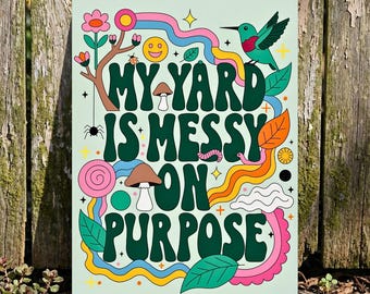 May include: A rectangular metal sign with the text "MY YARD IS MESSY ON PURPOSE" in dark green, surrounded by colorful illustrations of a hummingbird, flowers, mushrooms, and a rainbow. The background is a light green color.