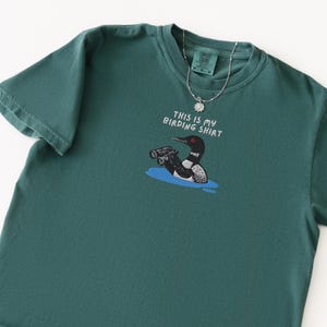 May include: Teal-colored t-shirt with a graphic of a loon wearing binoculars and the text "THIS IS MY BIRDING SHIRT." A silver necklace with a sun-shaped pendant is draped over the shirt. The shirt is made of a soft material.
