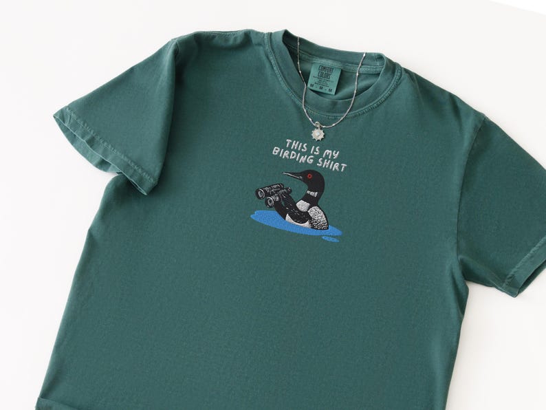 May include: Teal-colored t-shirt with a graphic of a loon wearing binoculars and the text "THIS IS MY BIRDING SHIRT." A silver necklace with a sun-shaped pendant is draped over the shirt. The shirt is made of a soft material.
