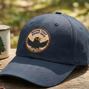 May include: Navy blue baseball cap with a circular patch featuring a black crow and the text "Gimme That Shiny Shit." The patch has a tan background with colorful coins at the bottom. The cap has a curved brim and a button on top.
