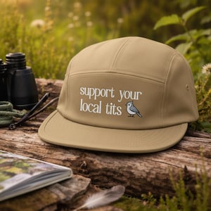 May include: A tan five-panel cap with a flat brim. The cap features the text "support your local tits" in white, along with a small illustration of a bird. The cap is resting on a wooden surface, with binoculars and a notebook in the background.