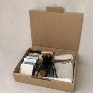 Peut inclure: Une boîte en carton brun contenant un assortiment d'échantillons de matériaux. La collection comprend plusieurs carreaux carrés dans des tons de gris, blanc et beige, un sac à cordon en organza noir avec des étiquettes circulaires dorées.