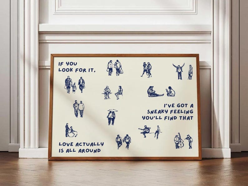 May include: A framed art print with a cream-colored background and a brown wooden frame. The print features blue line drawings of people and text that reads "IF YOU LOOK FOR IT," "LOVE ACTUALLY IS ALL AROUND," and "I'VE GOT A SNEAKY FEELING YOU'LL FIND THAT."