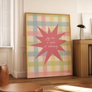 May include: A framed wall art print featuring a pastel plaid background in shades of blue, pink, yellow, and green. Centered on the print is a large, dark pink starburst shape containing the white handwritten text, joy has a habit of returning.