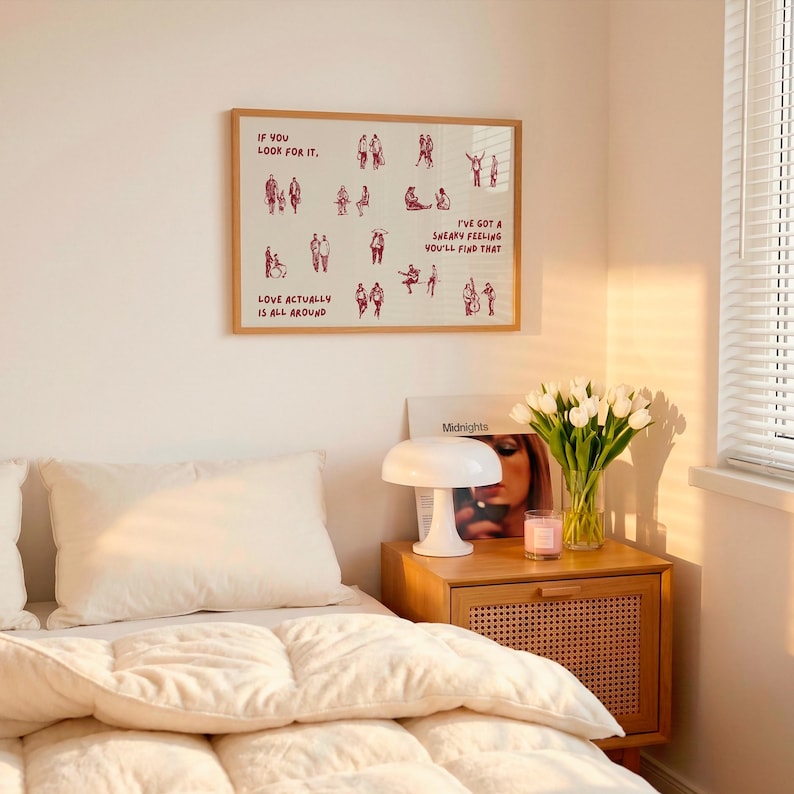 May include: A framed print with the text "IF YOU LOOK FOR IT, LOVE ACTUALLY IS ALL AROUND, I'VE GOT A SNEAKY FEELING YOU'LL FIND THAT" and illustrations of people. A white lamp, a vase of white tulips, and a wooden nightstand are also visible.