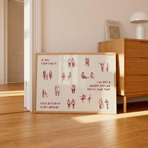 May include: A framed art print with a cream background and a wooden frame. The print features red illustrations of people in various poses, with text that reads "If you look for it," "Love actually is all around," and "I've got a sneaky feeling you'll find that."