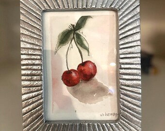 Könnte beinhalten: Ein kleiner, gerahmter Aquarellmalerei von zwei roten Kirschen mit grünen Blättern. Das Gemälde befindet sich in einem silberfarbenen Rahmen mit einem strukturierten, geriffelten Design. Die Signatur des Künstlers, "v. harvey", ist unten rechts sichtbar.