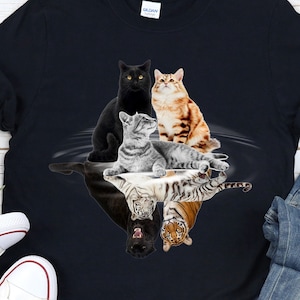 Könnte beinhalten: Ein schwarzes T-Shirt mit einem Aufdruck von vier Katzen und ihren Spiegelbildern im Wasser. Die Katzen sind eine schwarze Katze, eine graue Tabby, eine braune Tabby und ein Tiger. Der Aufdruck hat eine kreisförmige Form.