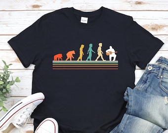 May include: A black t-shirt featuring a vintage-style graphic depicting the evolution of humans, culminating in a person playing a guitar. The graphic is in shades of orange, yellow, green, and blue.