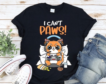 Może przedstawiać: Czarny T-shirt z nadrukiem kreskówkowego pomarańczowego kota w słuchawkach i trzymającego kontroler do gier wideo. Na koszulce widnieje napis "I can't paws!"