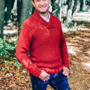 May include: A man wearing a red collared sweater and blue jeans stands in a forest setting. He is smiling and looking at the camera.