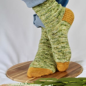 May include: A pair of green and yellow patterned socks with a yellow toe and heel. The socks are made of a soft, textured yarn and are shown on a wooden surface.