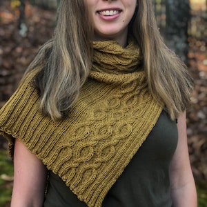 May include: A woman wearing a mustard yellow knitted shawl with a cable knit pattern. The shawl is draped around her neck and shoulders.