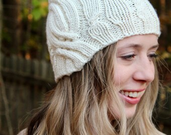 May include: A white knitted hat with a cable knit pattern. The hat is worn by a person with long blonde hair.