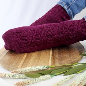 Op de afbeelding: Een paar bordeauxrode gebreide sokken met een kabelbreipatroon. De sokken liggen op een houten oppervlak met een takje gedroogde bloemen.
