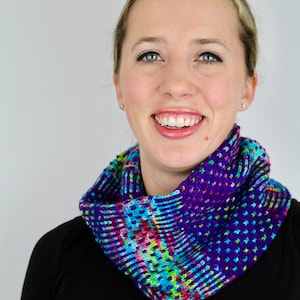 May include: A woman wearing a black shirt and a colorful knitted cowl neck scarf. The scarf has a purple, blue, green, pink, and white pattern.