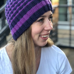 May include: A purple and blue striped knitted beanie hat. The hat is worn by a person with long blonde hair.