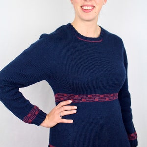 May include: A navy blue knitted dress with long sleeves and a round neckline. The dress has a burgundy geometric pattern around the waist, cuffs, and hem. A pocket is visible on the left side. The model is smiling.